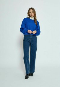 Modra pletena jopica z balonastimi rokavi, v kombinaciji z visokim pasom temnimi denim hlačami in črnimi gležnjakami. Stoji ob svetli podlagi.