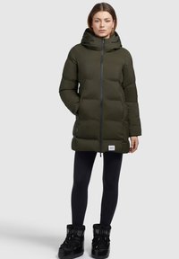 Olivgrüner, gepolsterter Mantel mit Kapuze, Frontreißverschluss und zwei Seitentaschen. Getragen mit schwarzen Leggings und schwarzen Schneestiefeln.