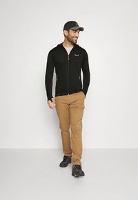 Mustast lukuga jakk oranžide detailidega, beežid töörõivad ja mustad kingad; peas tume müts. Seistes neutraalsel taustal.