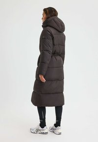 Mörk, vadderad pufferjacka med huva, har sidofickor, en åtdragen midja och en knälång design. Bärs över svarta leggings och skor.