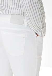 Weiße Denim-Jeans mit glatter Textur verfügen über eine Gesäßtasche und ein graues, gebrandetes Lederpatch. Ein gestreiftes Hemd blitzt von der Seite hervor.