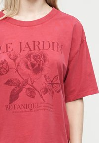 Κόκκινο μπλουζάκι μεγάλου μεγέθους με βοτανικό σχέδιο που περιλαμβάνει τριαντάφυλλα, πεταλούδες και το κείμενο «Le Jardin Botanique» στο μπροστινό μέρος.