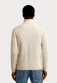 Beiger Rippenstrickpullover mit hohem Kragen, der ein strukturiertes Muster und kontrastierende Sprenkel aufweist, kombiniert mit blauen Denim-Jeans.