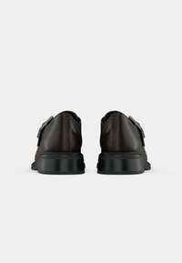 Chaussures habillées en cuir marron avec semelles en caoutchouc noir, vues de dos sur un fond gris clair uni.