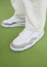 Hvite sneakers med en sølvglitrende seksjon langs siden, snøring og en strukturert hvit såle på en grønn overflate.