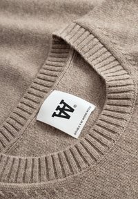 Beige strikket sweater med rund hals, der har ribbet manchetter og kant. Har et mærke med branding syet ind i halsudskæringen. Glat tekstur.