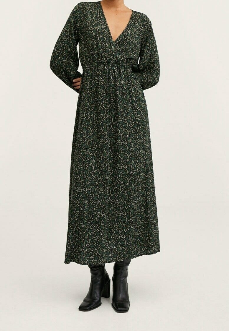 Femme vêtue d'une robe longue à manches longues à imprimé floral vert foncé avec un décolleté en V profond et des bottines noires à talons, mains sur les hanches.