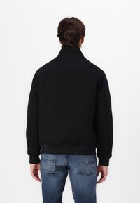 Svart hettegenser med høy krage og raglanermer, laget av et glatt stoff. Sammen med blå denimjeans. Enkel design, uten mønstre.