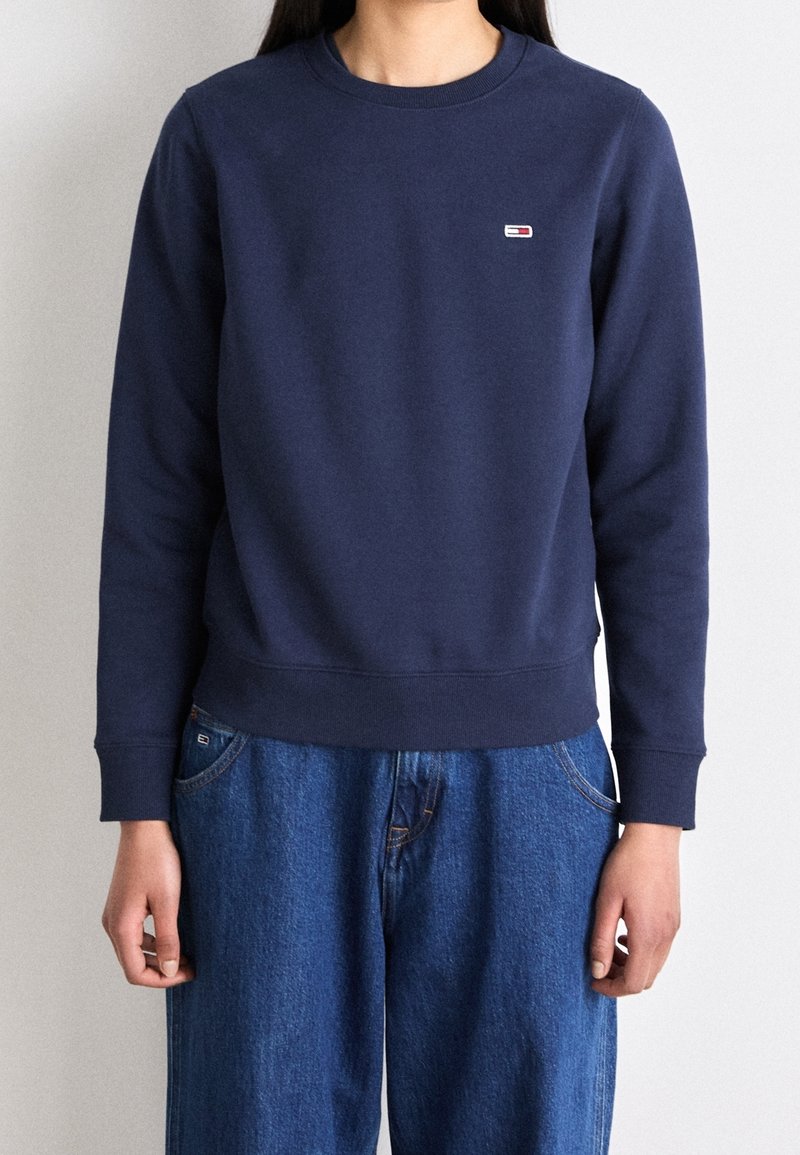 Person som bär en navyblå långärmad sweatshirt med en liten logotyp på bröstet och avslappnade blå jeans framför en neutral bakgrund.