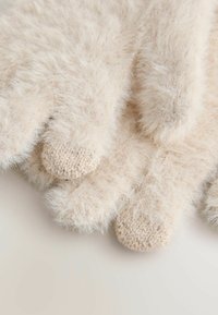 Mjuka, krämfärgade, fluffiga stickade handskar med texturerade fingertoppar, ordnade överlappande på en ljus yta.