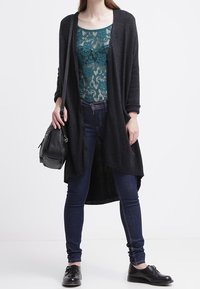 Svart lång cardigan över en teal spets-topp, kombinerad med mörka slim fit-jeans och svarta skor. Modell som håller en svart handväska. Platta, släta texturer.