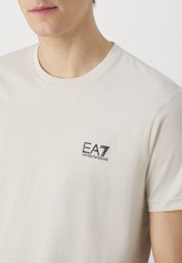 Krémové bavlněné tričko s malým černým logem EA7 Emporio Armani na levé části hrudi. Krátké rukávy a kulatý výstřih.