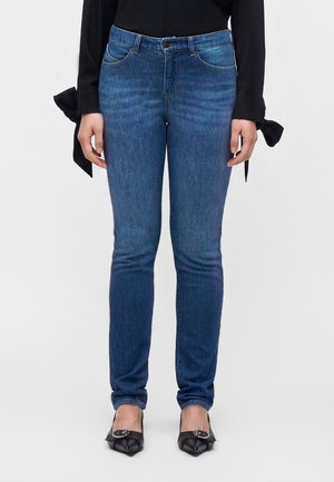 Person trägt eng anliegende blaue Jeans, schwarzes langärmeliges Oberteil mit großen seitlichen Schleifen und schwarze spitze Ballerinas mit silbernen Schnallen, steht aufrecht.