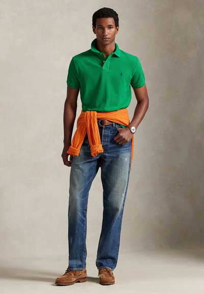 Πράσινο πόλο πουκάμισο με μπλε λογότυπο, πορτοκαλί πλεκτό πουλόβερ δεμένο γύρω από τη μέση, ανοιχτό μπλε τζιν, καφέ παπούτσια, ουδέτερο φόντο.