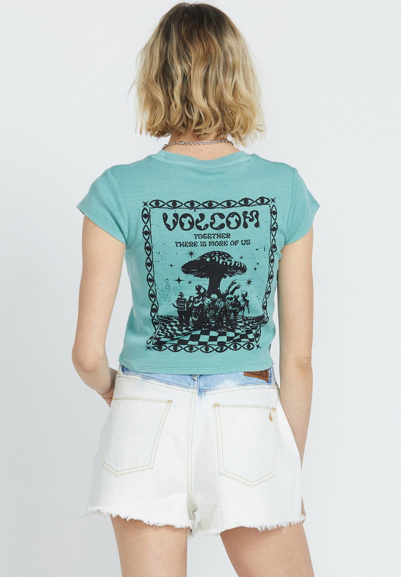 Teal T-shirt met korte mouwen, voorzien van een zwart grafisch ontwerp met tekst en beelden op de achterkant, gecombineerd met witte denim shorts.