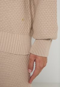 Beige gestrickter Pullover und Rock mit gerippten Bündchen und Saum, zeigt strukturierten Stoff und einen Teil einer Hand an der Seite.