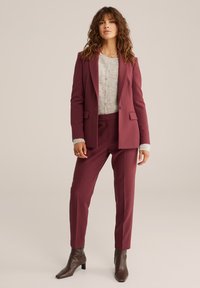 Burgundy skräddarsydd kostym med en kavaj som har spetsiga slag och matchande smalare byxor. Kombinerat med en ljus, strukturerad tröja. Bruna ankelboots.