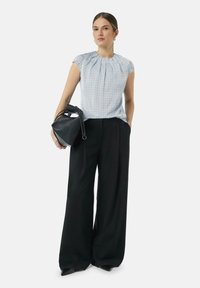 Weiße und blaue gemusterte Kurzarmbluse, kombiniert mit hochtaillierten, schwarzen weiten Hosen. Das Model hält eine kleine schwarze Tasche.