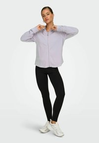 Jasnofioletowa kurtka zapinana na zamek w miękkim materiale, zestawiona z czarnymi legginsami i białymi sneakersami. Posiada swobodny krój i minimalne detale projektu.