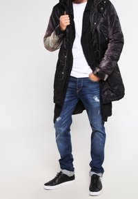 Dlouhá černá péřová bunda s kapucí, šedé rukávy, zapínání na zip, v kombinaci s modrými sepranými džíny a černými teniskami s bílou podrážkou.