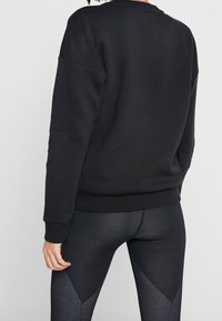 Felpa nera realizzata in un tessuto morbido, con maniche lunghe, scollo rotondo e polsini a costine. Abbinata a leggings neri con texture.