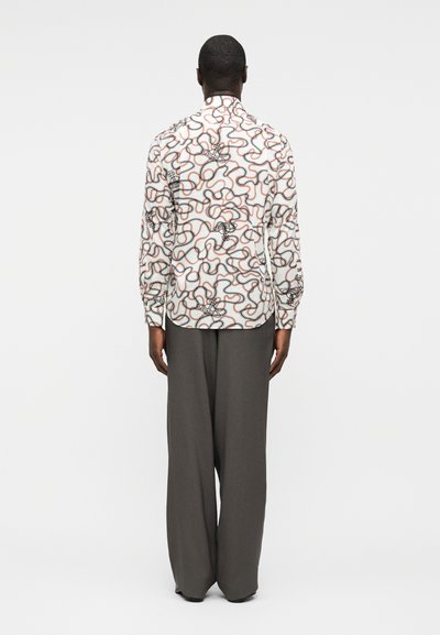 Άνδρας που στέκεται με την πλάτη στραμμένη, φοράει λευκό πουκάμισο με μακριά μανίκια με μαύρα και πορτοκαλί αφηρημένα σχέδια γραμμών και φαρδιά γκρι παντελόνια.