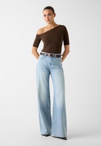 Brązowa prążkowana bluzka na jedno ramię oraz jasnoniebieskie jeansy o szerokich nogawkach z brązowym paskiem. Jeansy mają efekt sprania i wysoki stan.