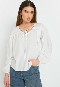 Hvid bluse med en afslappet pasform, puffærmer og snørelukning ved halsen. Har flæse detaljer langs skuldre og manchetter. Samlet med blå jeans.