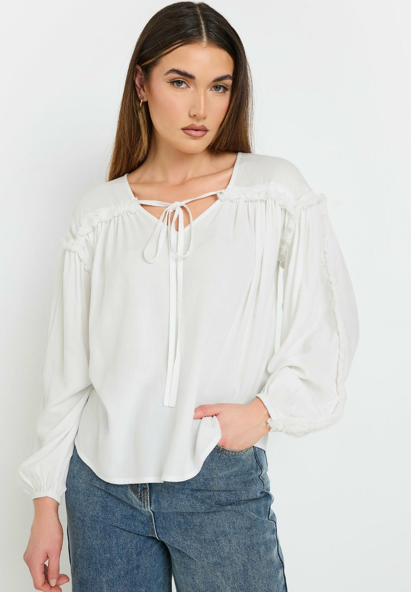 Hvid bluse med en afslappet pasform, puffærmer og snørelukning ved halsen. Har flæse detaljer langs skuldre og manchetter. Samlet med blå jeans.