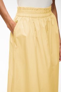 Personne portant une jupe jaune pâle, taille haute avec une ceinture élastique, main gauche dans la poche, vue de la taille au milieu de la cuisse.