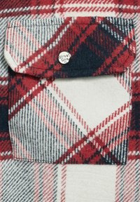 Rood, marineblauw en witte geruite stof met een vierkante zak en een zilveren drukknoop met het label "Street One." Zachte textuur, wolachtig materiaal.