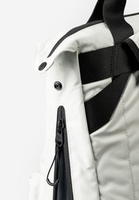 Close-up of white backpack corner with black zipper, black pull tab, and adjustable black strap on a white background.