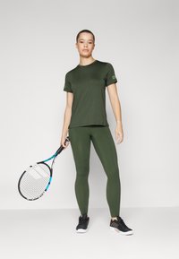 Zöld sportos felső és leggings sima textúrával, fekete sportcipővel párosítva, teniszkütővel a semleges háttér előtt.