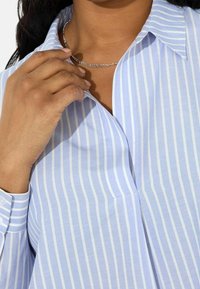 Camicia a righe azzurro chiaro con polsini abbottonati, tessuto morbido, colletto appuntito e una patta poco profonda; mano appoggiata sul colletto.