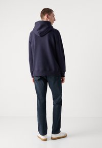Sudadera con capucha en color navy, con puños y dobladillo acanalados, combinada con jeans oscuros y zapatillas blancas, vista desde la parte de atrás.