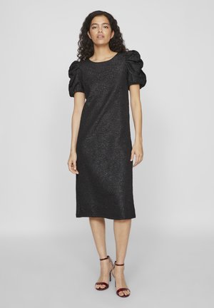 Robe noire midi avec un motif floral texturé, décolleté rond et manches bouffantes. Associée à des sandales à talons hauts bordeaux.