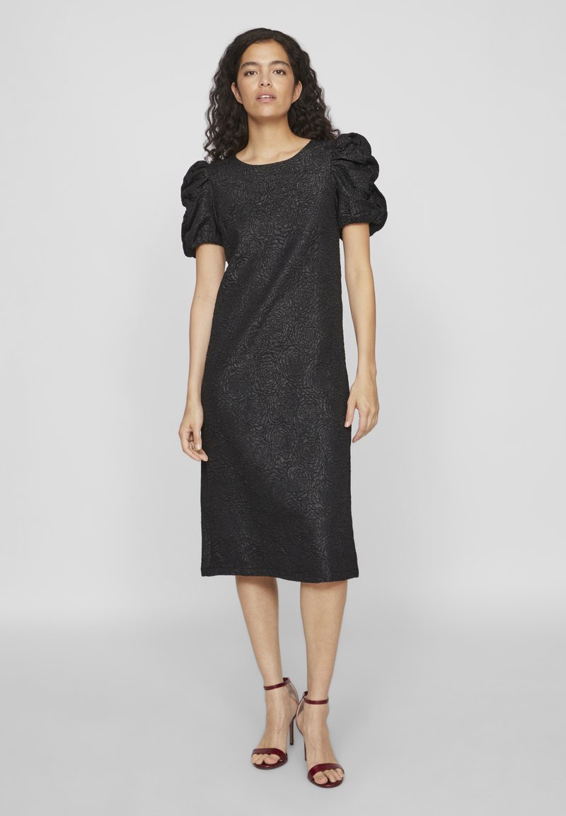 Γυναίκα που φοράει ένα μαύρο φορεμά πάνω από το γόνατο με φουσκωτά κοντά μανίκια και φλοράλ υφή, συνδυασμένο με κόκκινες ψηλοτάκουνες σάντες με λουριά, στέκεται σε γκρίζο φόντο.