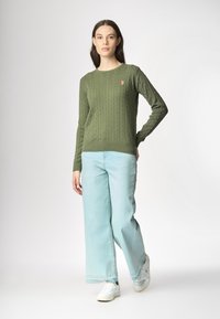 Una giovane donna sta in piedi con sicurezza, indossando un maglione verde a trecce e jeans a gamba larga di un blu chiaro, con sneakers bianche.