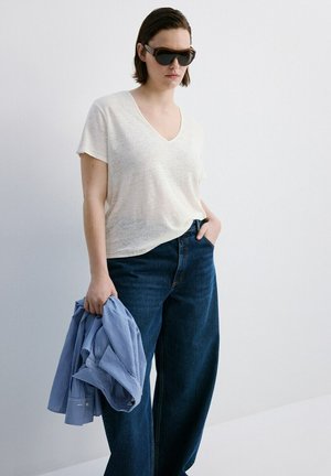 Femeie purtând ochelari de soare închiși la culoare, tricou alb cu decolteu în V, blugi albaștri cu picioare largi, ținând o cămașă albastră cu dungi într-o mână.