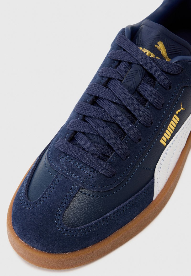 Zapatillas de deporte azul marino con materiales de cuero y ante, que cuentan con una suela de goma, cordones texturizados y una franja lateral blanca con branding dorado.