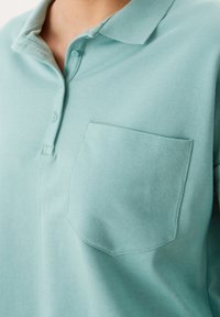 Polo de color verde claro hecho de una suave mezcla de algodón. Presenta un clásico cuello, una tapeta con tres botones y un solo bolsillo en el pecho.
