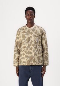 Tan camouflage quiltet jakke med rund krave, knaplukning, sidelommer og et lille logo på venstre bryst.