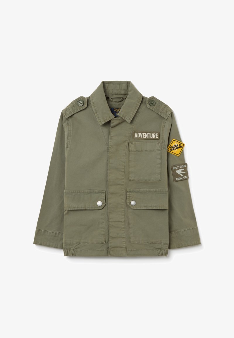 Veste vert olive avec un col, des poches avant et des épaulettes à boutons. Comprend des détails en patch : "AVENTURE !" et un triangle de sécurité.