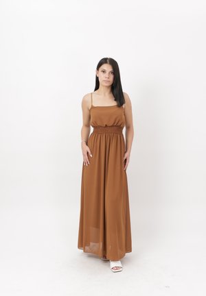 Femme aux cheveux lisses et foncés, portant une robe maxi marron sans manches et des chaussures blanches à bout ouvert, debout devant un fond blanc.