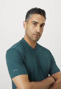Moški s kratkimi temnimi lasmi in sivimi prameni nosi teal kratkolasni športni majico, križanih rok na enobarvni beli podlagi.