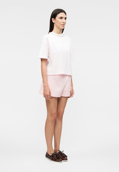 Γυναίκα στέκεται φορώντας ανοιχτόροζ μπλουζάκι με κοντά μανίκια και αντίστοιχα σόρτς με καφέ loafers με φούντες σε λευκό φόντο.