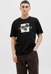 Camiseta negra de algodón con un gráfico frontal de siluetas de plantas sobre un cuadrado blanco, con el texto "ORIGINALS" en dorado. Manga corta.