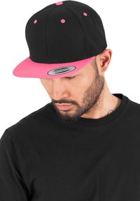 Černá snapback kšiltovka s růžovým plochým kšiltem a odpovídajícími akcenty. Na kšiltech je umístěna kulatá nášivka s logem. Nositá pod úhlem.