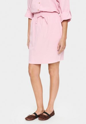 Jupe à cordon de serrage rose clair en tissu texturé, dotée d'une taille élastique et de poches latérales, associée à des chaussures plates marron.
