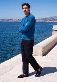 Man wearing blue sweater and black pants stands with arms crossed on waterfront promenade with sea and hills in background.